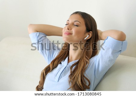 Chill out relax at home. Blissful young woman sitting listening to music with wireless earphones on her mobile phone at home as she relaxes with closed eyes on a sofa.
