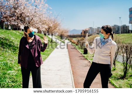 Two friends with protective masks greet with waving to each other.Alternative greeting during quarantine to avoid physical contact.Coronavirus COVID-19 disease protection.Social distancing practice