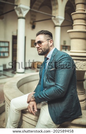 Handsome looking man in smart combination dressed in blue blazer and cream pants wearing sunglasses,sitting on the fountain, outside portrait