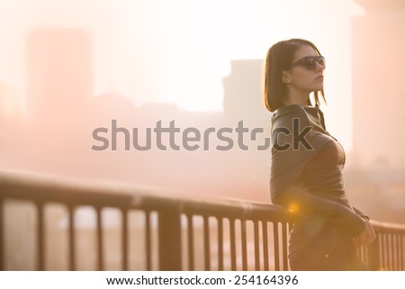 Rocker,punk woman.Young rock woman in black leather jacket casual standing with city background at sunset.Rock bad girl.Sexy attractive woman with punk rock fashion styling.Image toned and noise added