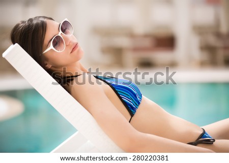 Sexy beautiful brunette woman with in bikini relaxing beside a swimming pool in summer on tropic island in hot weather on pool party.Woman relaxing in deck chair.Sunscreen,protection,summer body