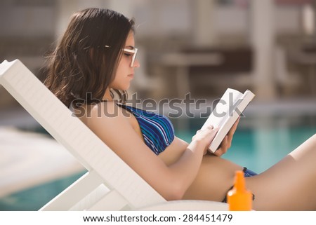 Sexy brunette woman in bikini relaxing near swimming pool on tropic island in hot weather on the seaside reading a book.Woman relaxing in deck chair.Sunscreen,protection,summer body,summer recreation