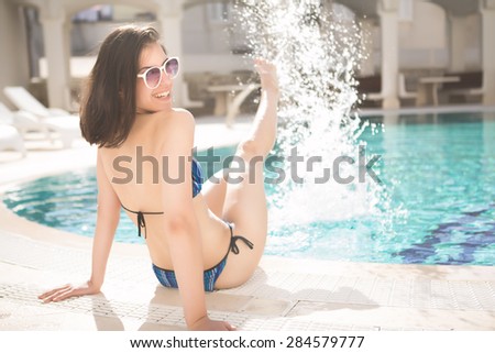 Young beautiful woman making water splash in sexy bikini at the pool.Enjoying summer.Young slim fit tanning woman having fun near poll and making water splash with her sexy legs. Vacation mood.