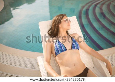 Sexy beautiful brunette woman with sunglasses in bikini relaxing near swimming pool in summer on tropic island in hot weather .Woman relaxing in deck chair.Sunscreen,protection,summer body