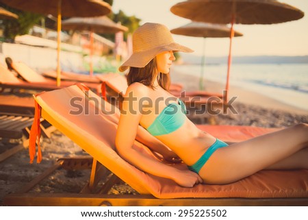 Beautiful woman sunbathing in a bikini on a beach at tropical travel resort,enjoying summer holidays.Young woman lying on sun lounger near the sea.Beach sunset and golden light.Woman in hat and bikini