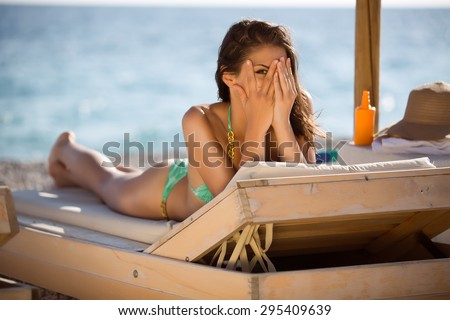 Shy woman peeking through covered face.Funny beautiful woman sunbathing in a bikini on a beach at travel resort,enjoying summer holidays.Young woman lying on sun lounger near the sea,having SPF creme