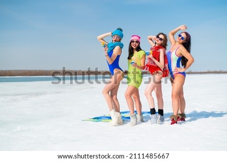 Four caucasian women in swimsuits pose and drink blue cocktails on a snowy beach.