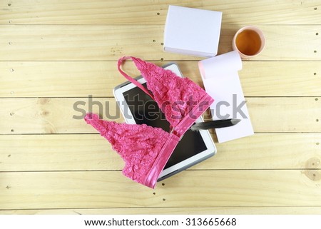 Tablet, pink bra, blank box, tea cup, feather pen and brown book are on wood table