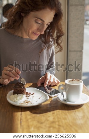 woman taking chocolate cake piece with spoon next to white small cup cappuccino coffee and touching mobile phone blank screen on light brown wooden table