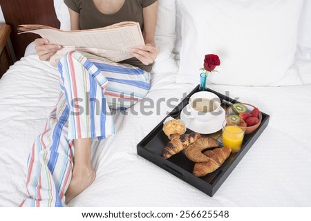 woman green shirt striped pajama pants sitting on white bed reading financial newspaper with breakfast tray croissants orange juice strawberry kiwi cupcake red rose flower