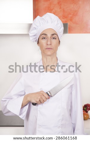 portrait of serious brunette chef woman with professional jacket and hat in white and orange kitchen saluting oath with big metal knife on chest