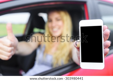 Happy blonde woman showing smartphone and showing thumb up out the window of a car. Focus on mobile phone.