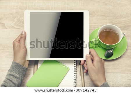 Tablet PC in the hands, a cup of tea and notebook on the desktop. Top view. Low contrast toned style.