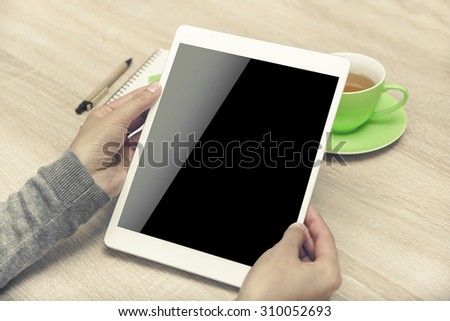 Tablet PC in the hands, a cup of tea and notebook on the desktop. Low contrast toned style.