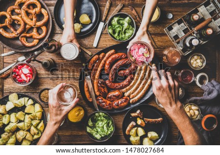 Flat-lay of Octoberfest party dinner table with grilled meat sausages, German pretzel pastry, potatoes, cucumber salad, sauces, beers and peoples hands with food over dark wooden background, top view Foto d'archivio © 