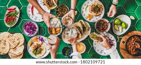 Friends home taco party. Flat-lay of Mexican traditional dishes Tacos with beef meat, corn tortillas , tomato salsa and peoples hands with glasses over green background, top view. Mexican cuisine