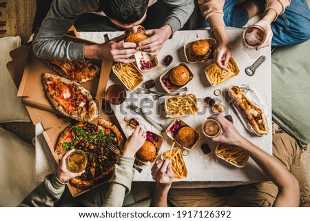 Lockdown fast food dinner from delivery service. Flat-lay of friends sitting and having beer quarantine party with burgers, french fries, sandwiches, pizza and salad over table background, top view
