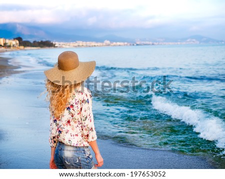 A young blondy girl in a sun-hat and flower-printed blouse staying at the sea coast and looking forward to the horizon
