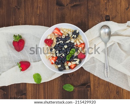 A bowl of oat porridge with fresh berries, honey, almond petals and leaves of mint over a piece beige linen fabric on a dark wooden background