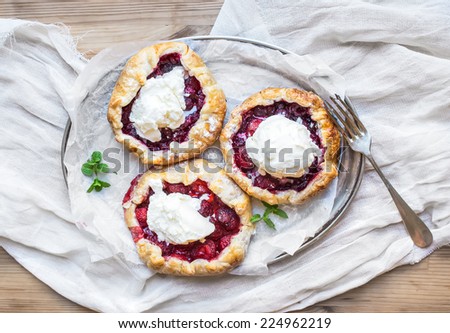 Rustic small galette pies with fresh berries and vanilla ice-cream on silver dish over a piece of white linen fabric and wooden background