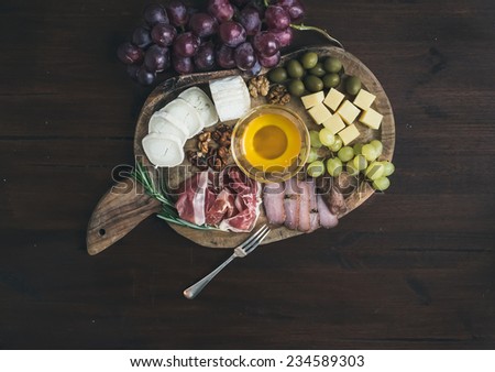 Wine appetizers set: meat and cheese selection, honey, grapes, walnuts and olives on a rustic wooden board over a dark wood background. Top view