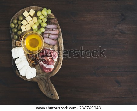 Wine appetizers set: meat and cheese selection, honey, grapes, walnuts and olives on a rustic wooden board over a dark wood background with a copy space. Top view