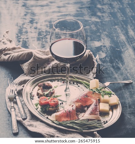Wine appetizer set. Glass of red wine, vintage dinnerware, brushetta with cherry, dried tomatoes, arugula, parmesan, smoked meat on silver tray over rustic grunge surface. Closeup