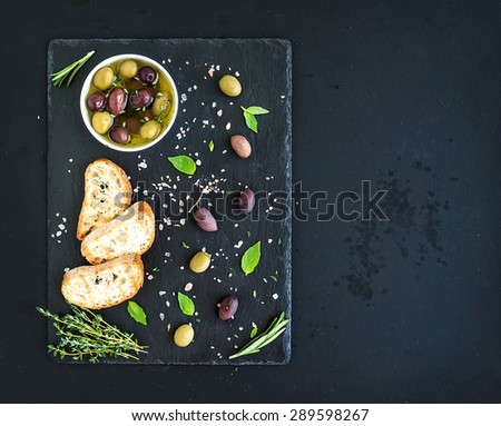Mediterranean olives with herbs and ciabatta slices on black slate tray over dark grunge background, top view, copy space