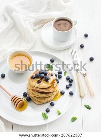 Breakfast set. Blueberry pancakes with fresh berries, honey, mint leaves and cup of coffee over white wooden backdrop