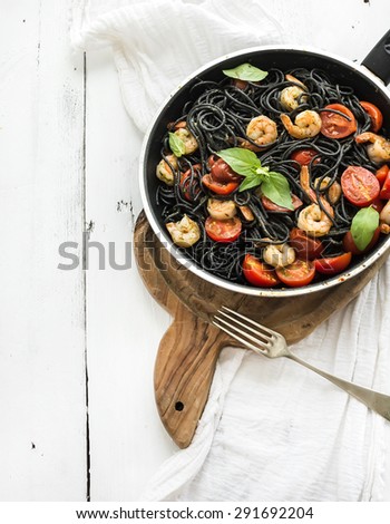 Black pasta spaghetti with shrimps, basil, pesto sauce and slow-roasted cherry-tomatoes in cooking pan on rustic chopping board over white wooden table, top view