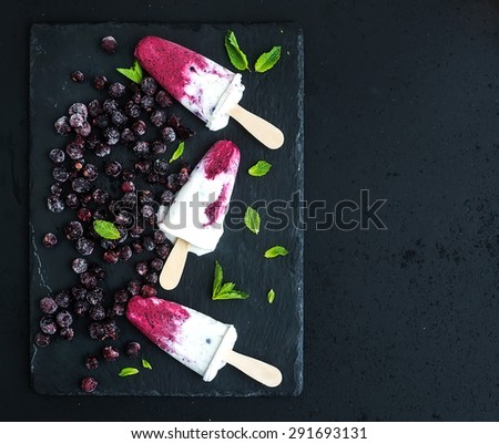 Black-currant and cream ice-creams or popsicles with frozen black-currant and mint on black slate tray over dark grunge backdrop, top view, copy space