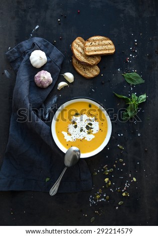 Pumpkin soup with cream, seeds, bread and fresh basil in rustic metal plate on grunge black background. Top view