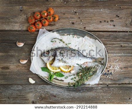 Roasted dorado or sea bream fish with vegetables, herbs and spices on silver tray over rustic wood backdrop, top view, copy space