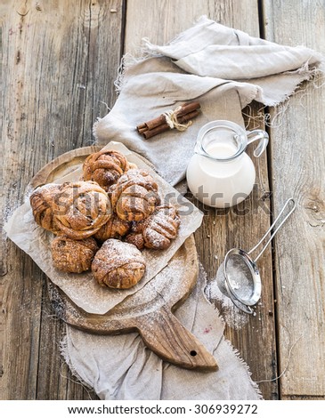 Cinnamon buns with sugar powder on a rustic wooden board, jug of milk on old wooden background