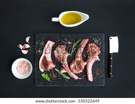 Raw lamb chops. Rack of Lamb with garlic, rosemary and spices on black slate tray, oil in a saucer, salt, dinnerware over dark rustic wood background