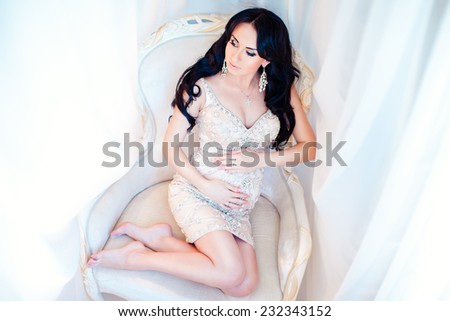 Beautiful slim pregnant woman in a luxury dress sitting on a chair and touches her tummy.