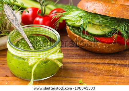 Green pesto in little jar. Vegan bagel and raw vegetables in the background. Selective focus on pesto.