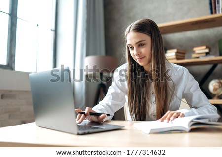 A beautiful caucasian female student is studying in college remotely. She is sitting at a table at home with a laptop and a notepad and concentrated is watching a video conference lesson
