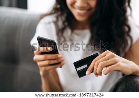 A credit card and smartphone in female hands. Joyful african american young woman in defocus looks at the phone screen, shopping online. Credit card and phone in the focus