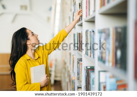 Caucasian female student stands in university library, looking for a book. Beautiful young woman in eyeglasses searching information for a project, learning for passing exams, education concept
