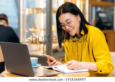 Modern Asian female student in stylish casual clothes, studying remotely using a laptop, listening to an online lesson, taking notes in a notebook, while sitting in a coffee shop. Online education