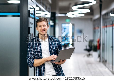 Caucasian guy freelancer with laptop. A successful guy dressed in stylish casual wear stands in a creative office, holds a laptop, looks and smiles friendly at the camera