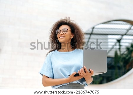 Photo of lovely elegant african american young woman with curly hair, in a blue shirt and glasses, standing outdoors, holding an open laptop, looks away, smiling happily, dreaming, thinking Foto stock © 