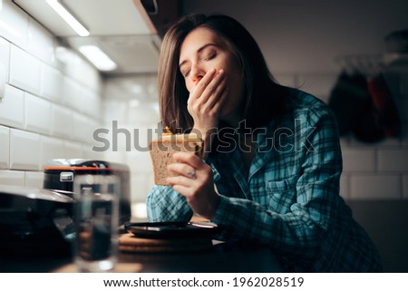 Sleepy Woman Holding a Sandwich in the Kitchen. Tired young person feeling hungry before going to sleep eating at night
