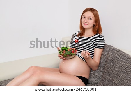 Healthy nutrition and pregnancy. Cheerful beautiful pregnant woman sitting on the sofa with a bowl full of vegetables.