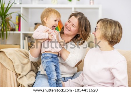 Two young women are sitting on the couch by the seyu at home in the living room and playing with their little daughter. Gay lesbian couple at home with a baby. Lesbian family 