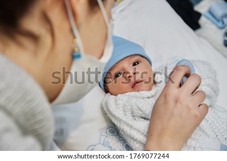 young redheaded mother with preventive health mask and lgbt bracelet, next to her newborn baby playing and laughing together on the bed, concept of family and newborn