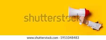 Hand holds a megaphone from a hole in the wall on a yellow background. Concept of hiring, advertising something. Banner.