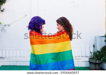 Two women with rainbow flag on a backyard. Couple Young lesbian girls holding a rainbow flag and hugging happily. A pair of lesbian women
