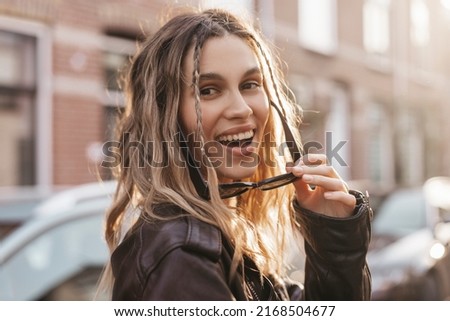 Pensive blonde woman in black leather jacket put off black glasses and turn around posing on street background. Outdoor shot of happy hippie lady with two thin braids and wave hair. Boho freedom style Foto stock © 